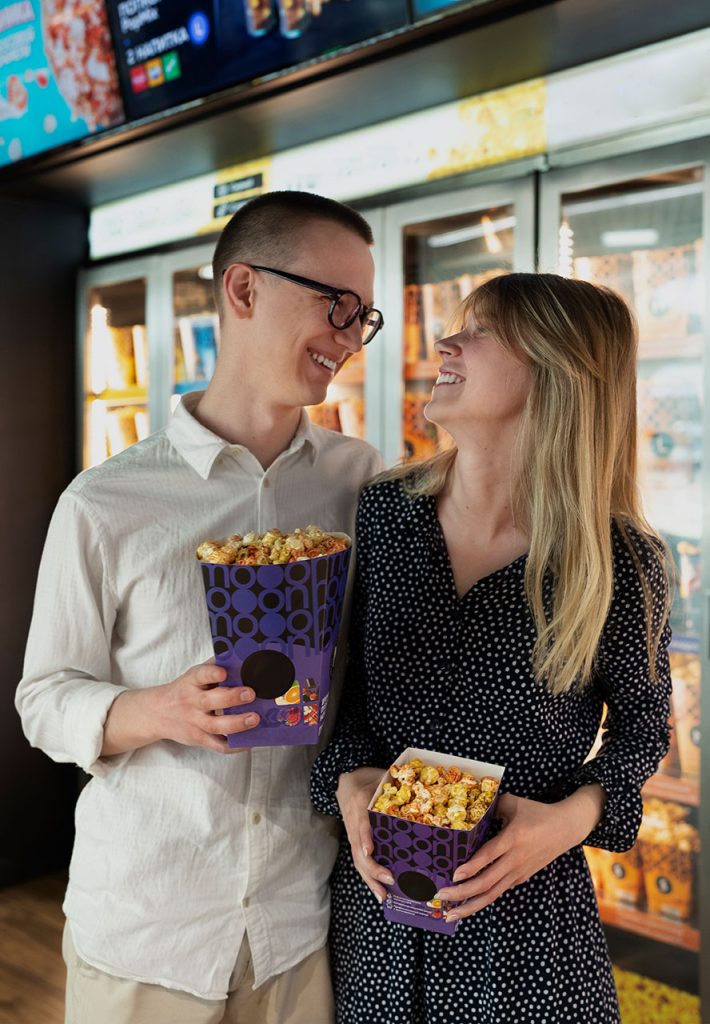 A smiling couple holding boxes of popcorn, standing in a cinema lobby, ready to enjoy their movie experience together.