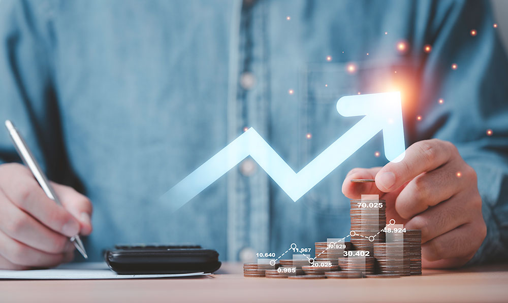 A person calculating finances with a virtual graph showing an upward trend over a stack of coins, symbolizing business growth, investment, and profit increase.